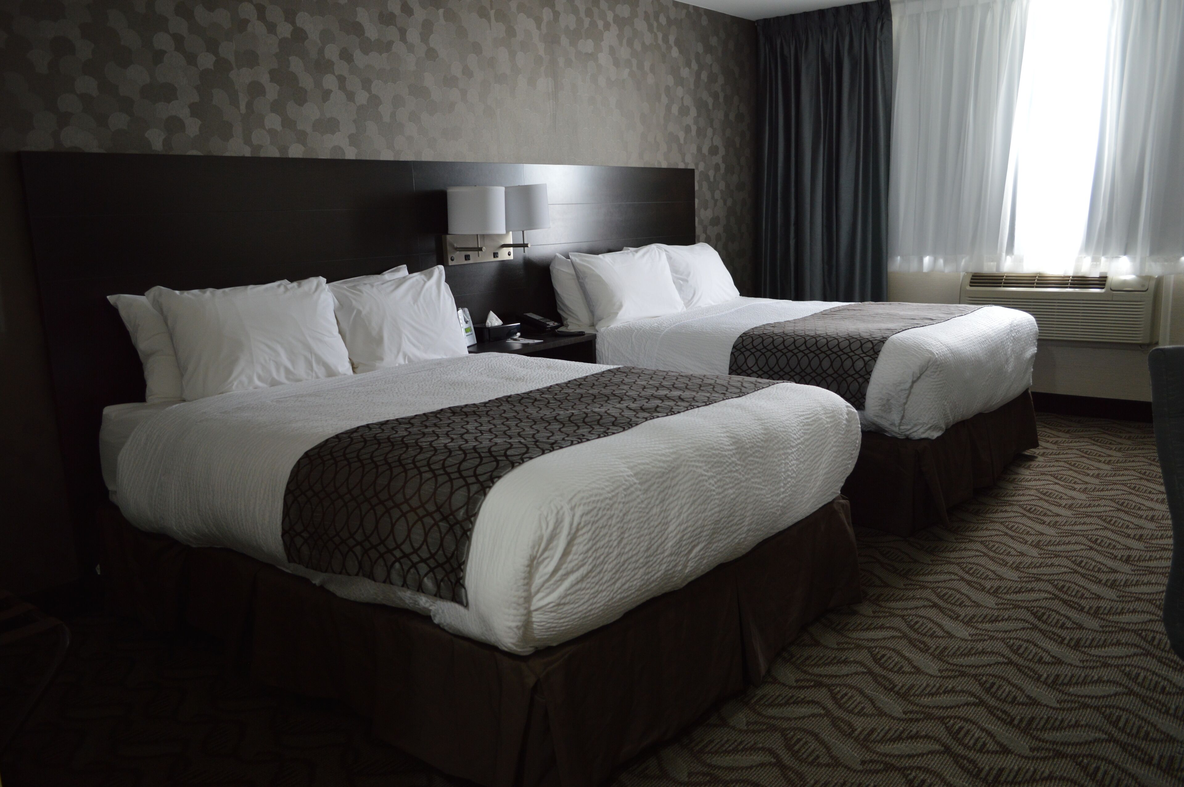 A hotel room with two beds, a headboard with a built-in TV, a bedside table, and a patterned carpet.