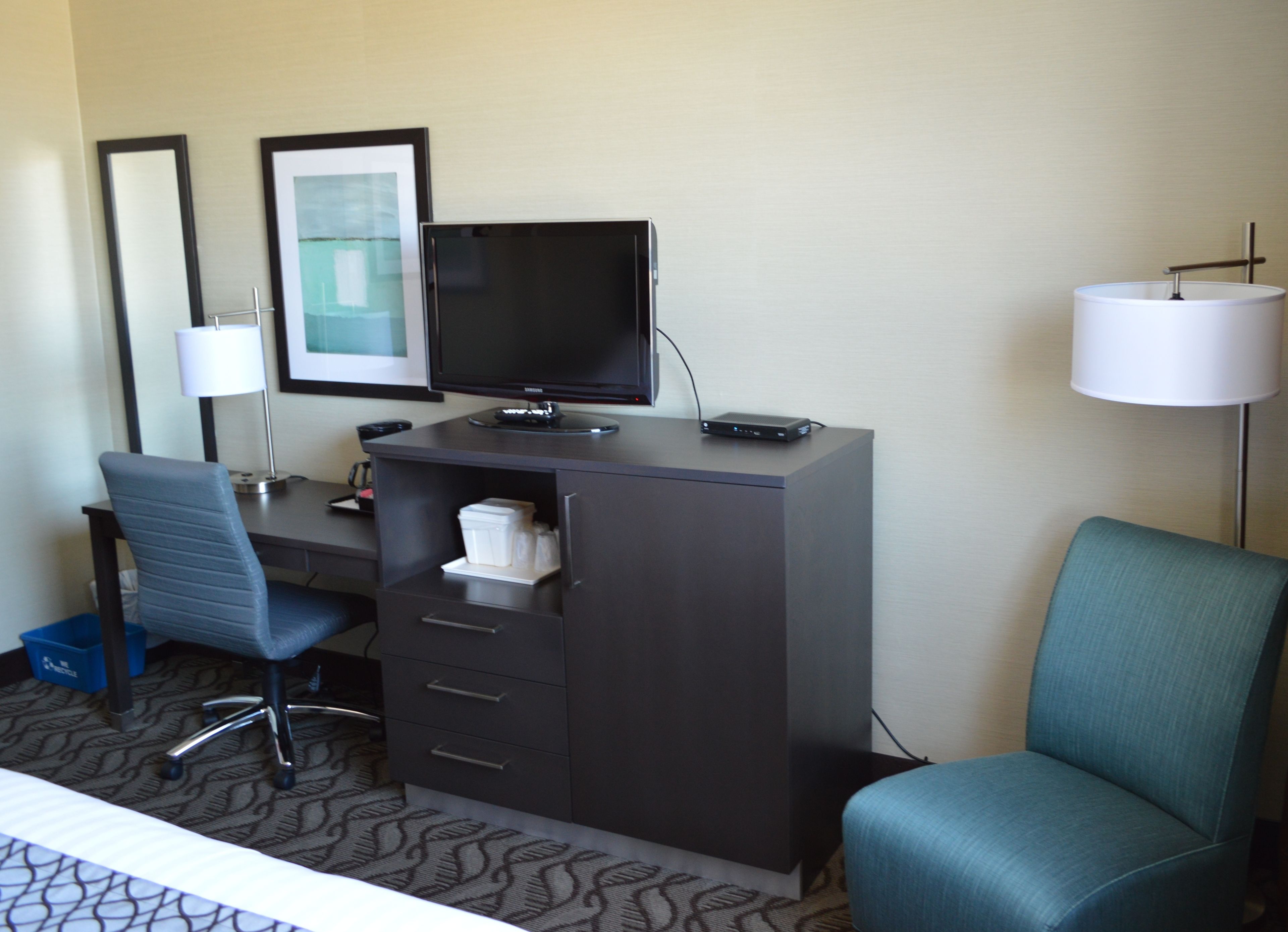 Una habitación de hotel con escritorio, computadora, silla, lámpara y cama.