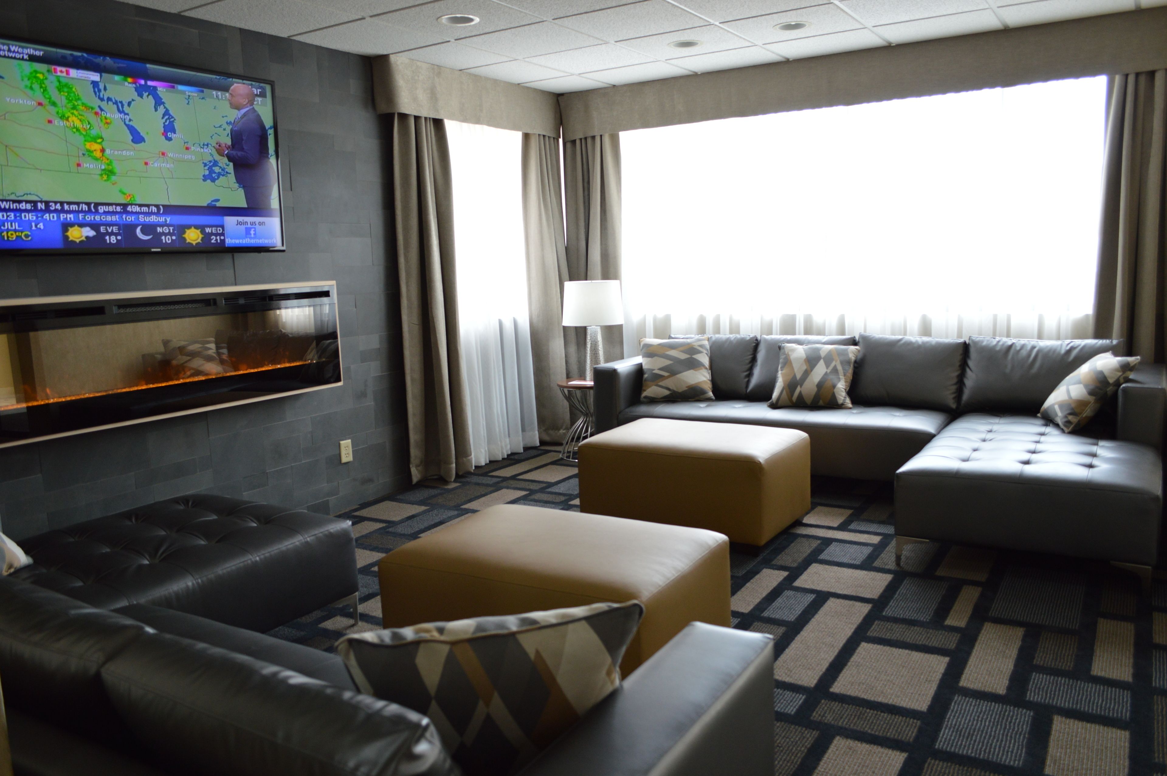 A modern hotel room with a large flat-screen TV displaying a weather forecast, a fireplace, a sofa set, and a large window with curtains.