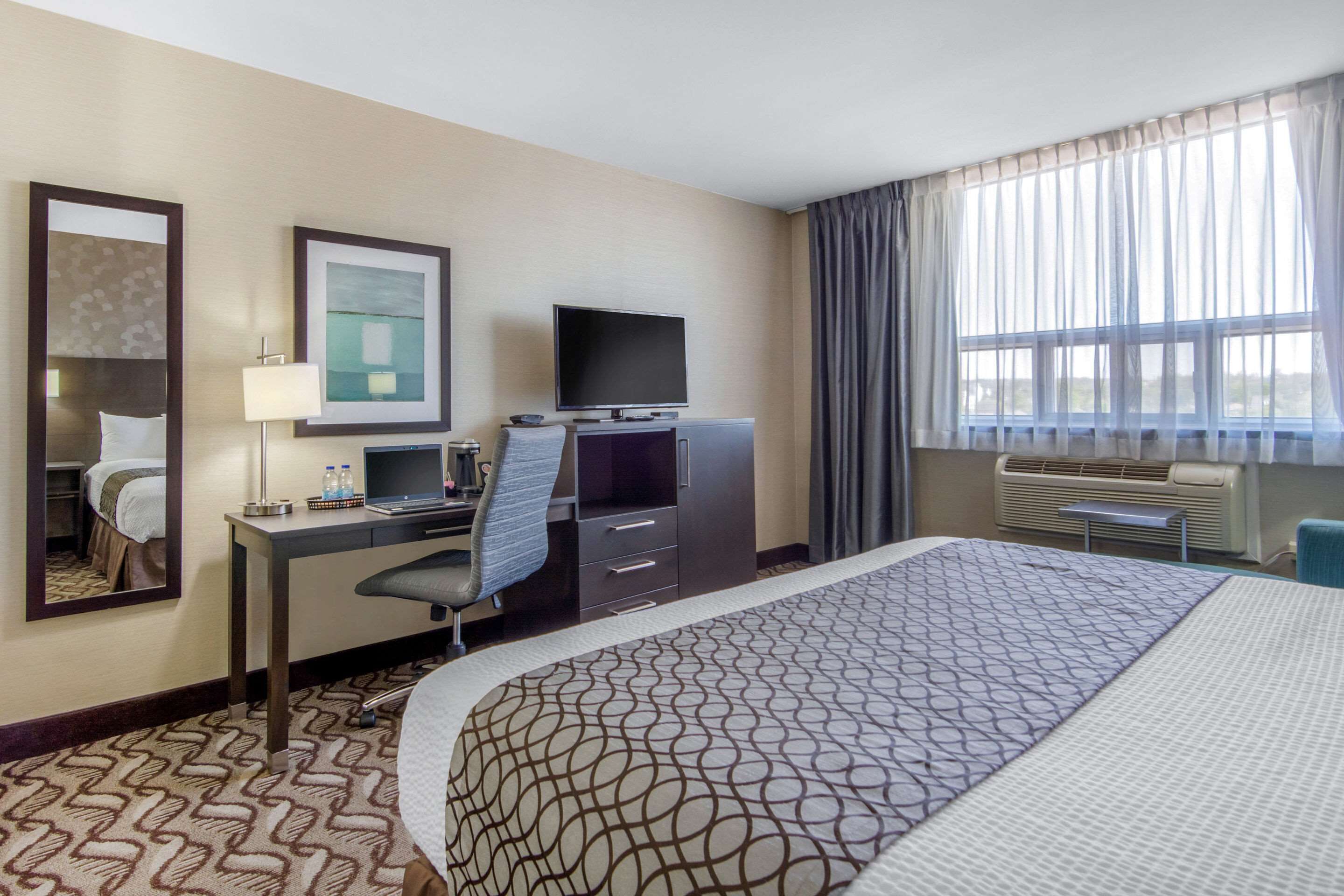 Habitación de hotel con una cama grande, un escritorio con ordenador, un televisor de pantalla plana, un espejo y una ventana con cortinas.