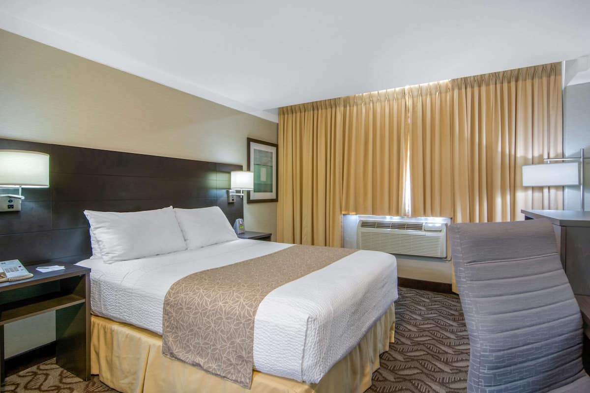 Ein Hotelzimmer mit einem Bett, einem Schreibtisch mit Lampe, einem Sessel, einem großen Fenster mit Vorhängen und einer Klimaanlage.