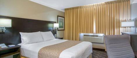 A hotel room with a bed, a desk with a lamp, a chair, a large window with curtains, and an air conditioning unit.