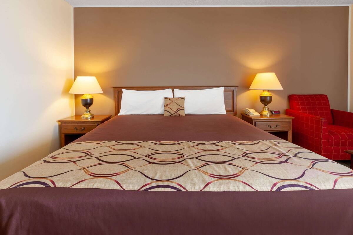 Habitación de hotel con una cama grande, dos mesitas de noche con lámparas, una silla roja y una colcha estampada.