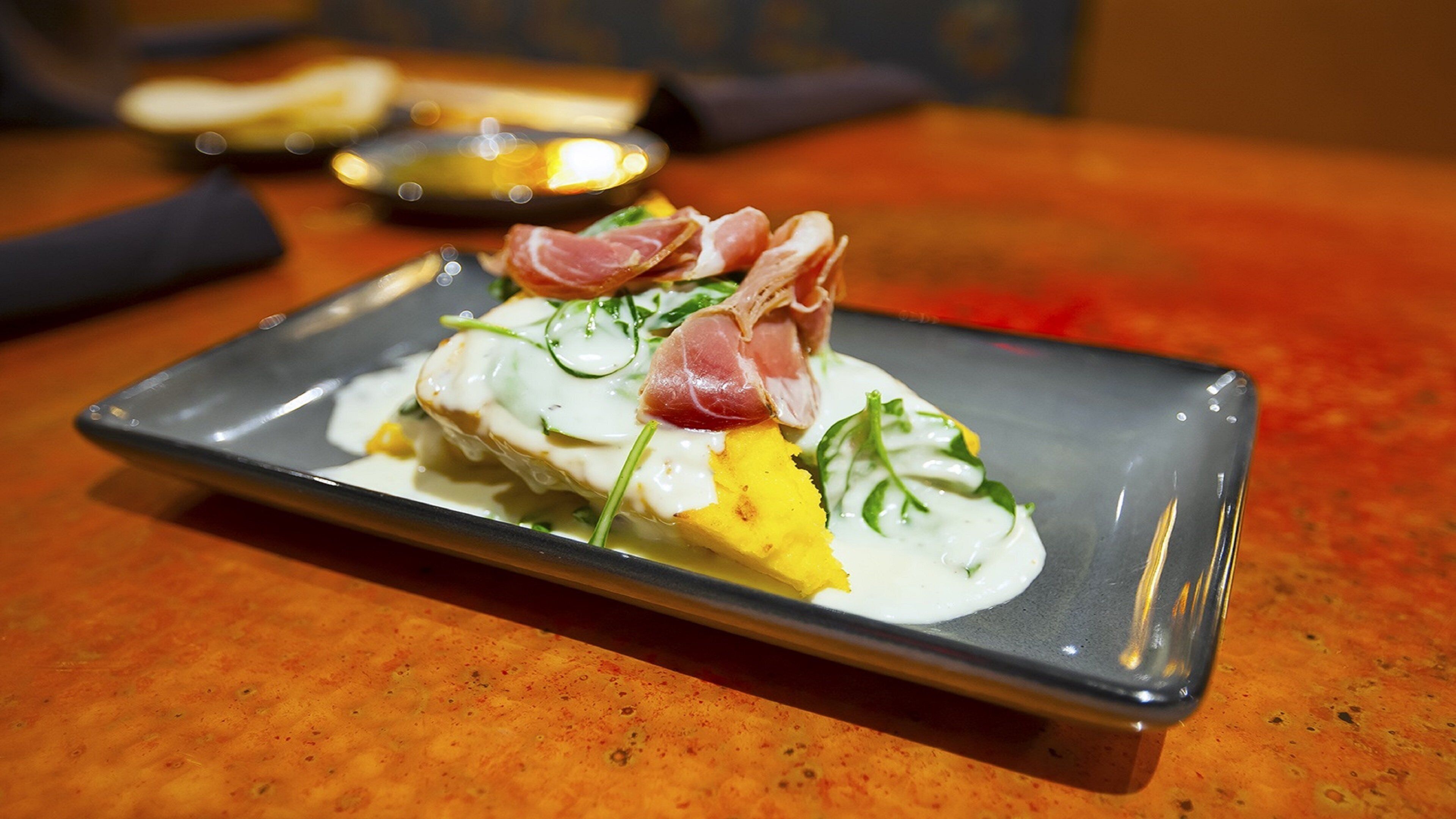 A rectangular plate with a white sauce, yellow food item, green garnish, and a slice of pink meat on a textured orange surface.