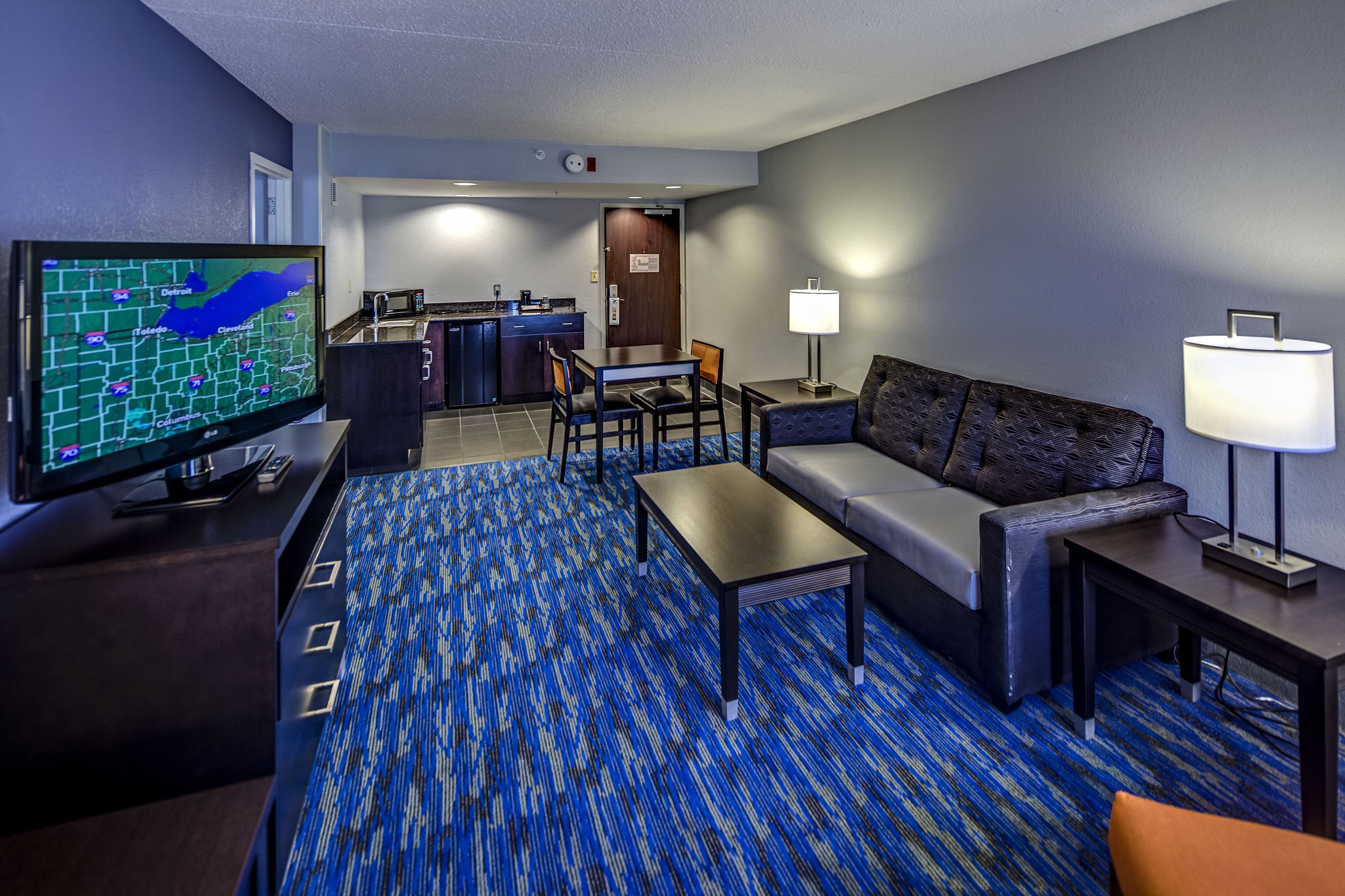 A hotel room with a television, a sofa, a coffee table, a lamp, and a carpeted floor.