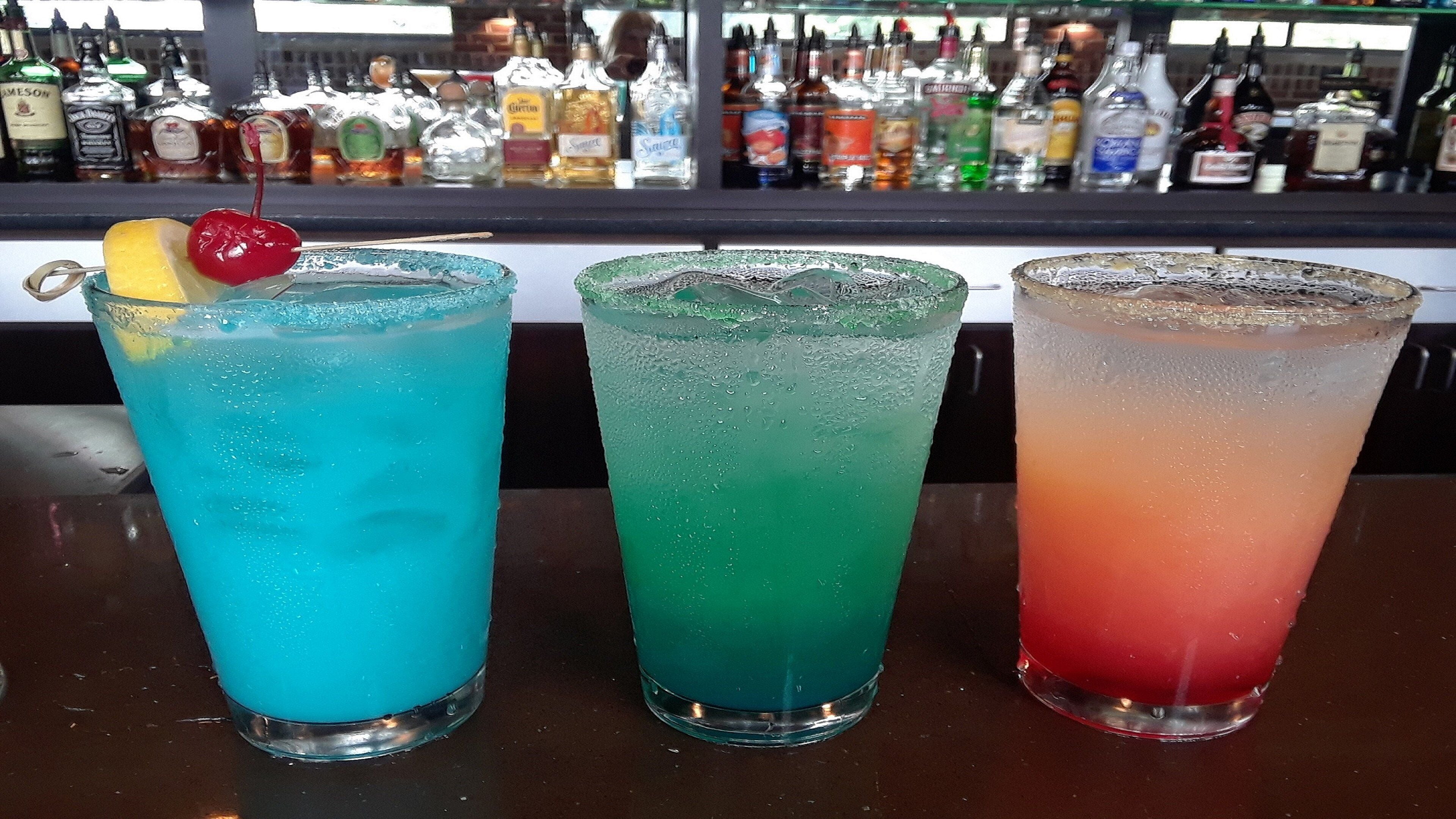 Three colorful drinks with a bar of various liquors in the background.