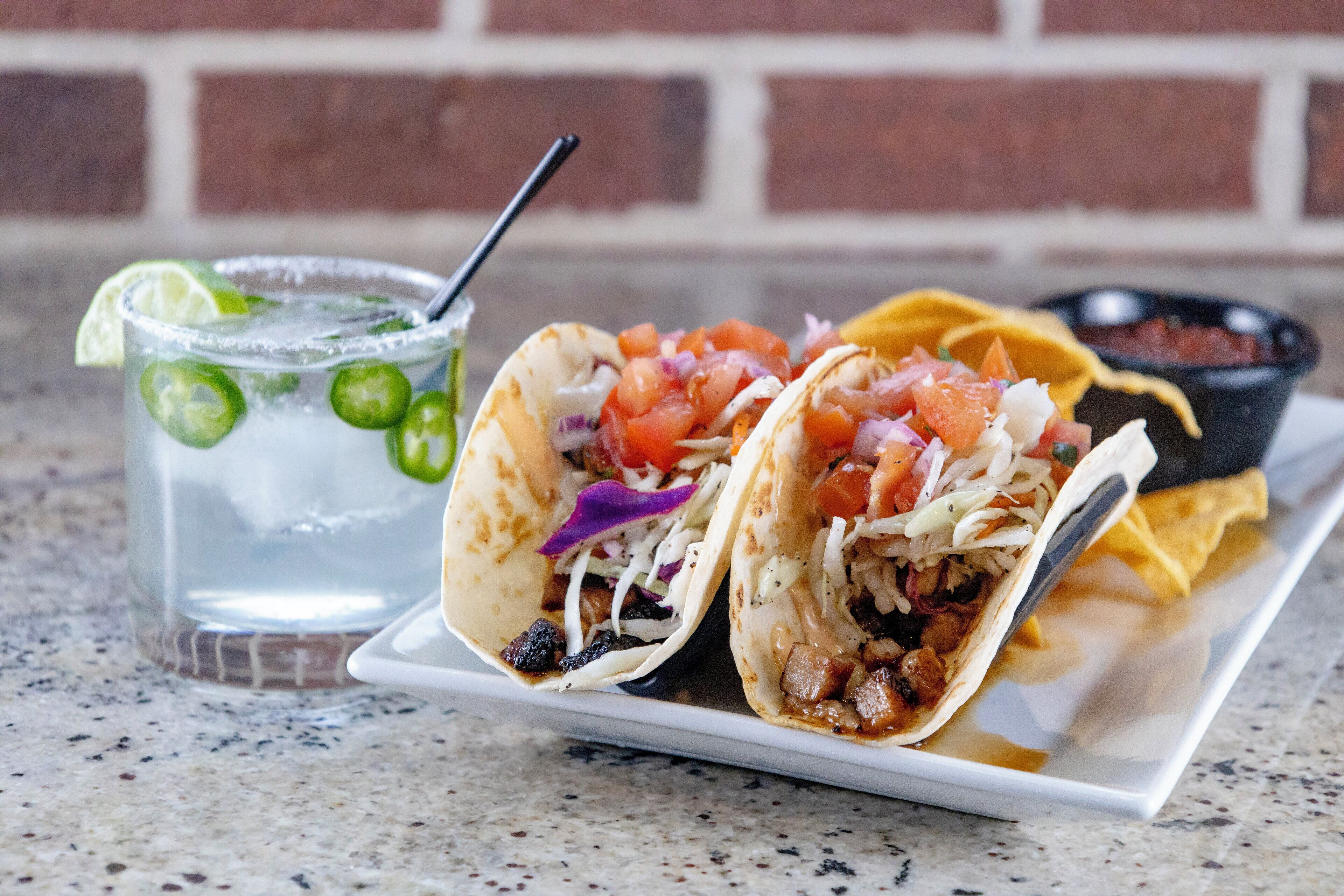 Dois tacos em um prato branco, acompanhados de um recipiente com salsa e um copo de bebida gelada com rodelas de limão.