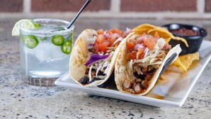 Deux tacos servis sur une assiette blanche, accompagnés d’une sauce salsa et d’un verre de boisson fraîche avec des rondelles de citron vert.