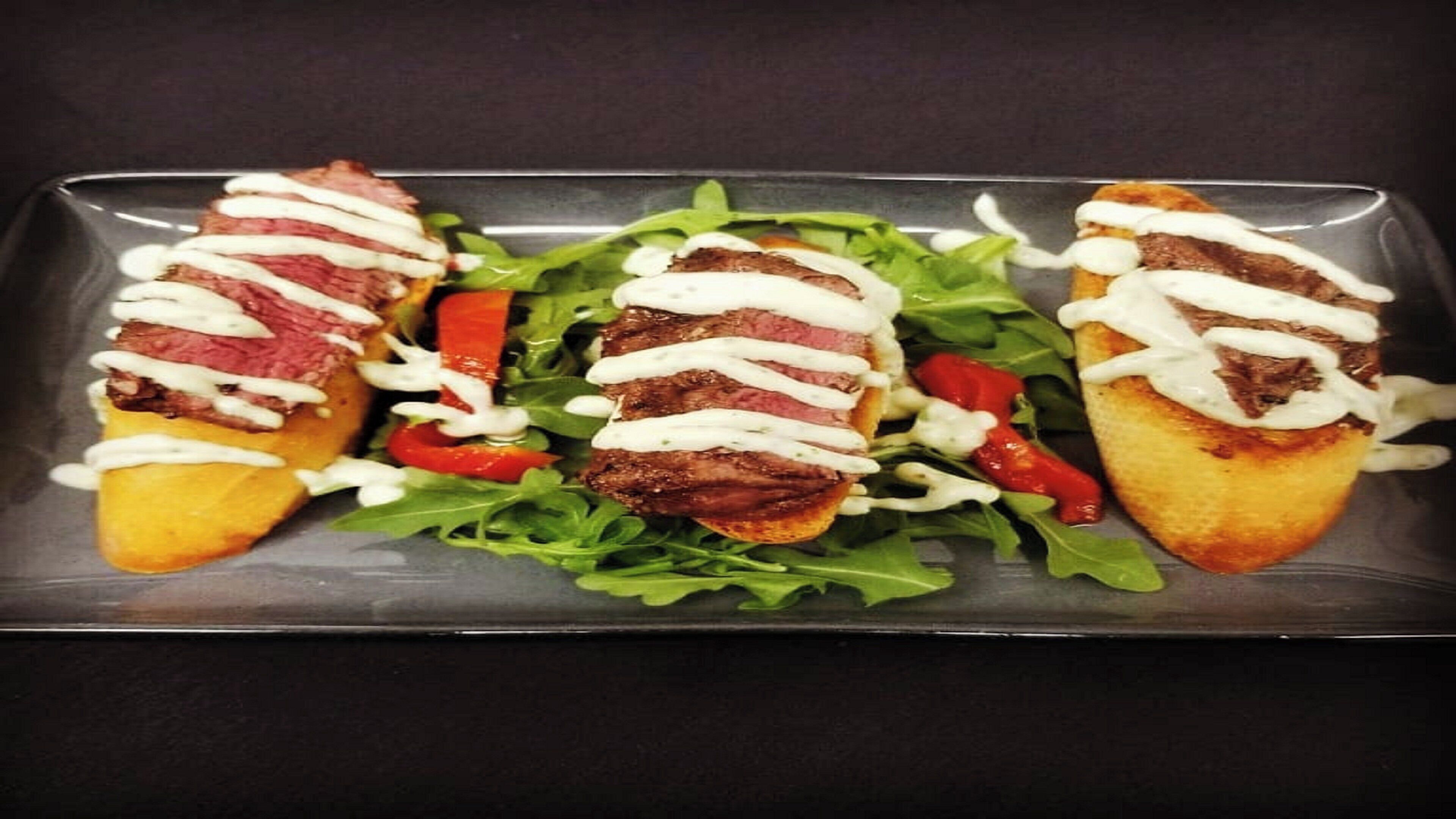 A rectangular plate with three grilled meat sandwiches topped with white sauce, accompanied by a bed of mixed greens and red bell peppers.