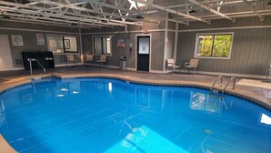Une piscine intérieure à l’eau d’un bleu limpide, entourée d’une terrasse carrelée. Des chaises et des tables sont disposées autour de la piscine, et un panneau indique qu’il est interdit de plonger.