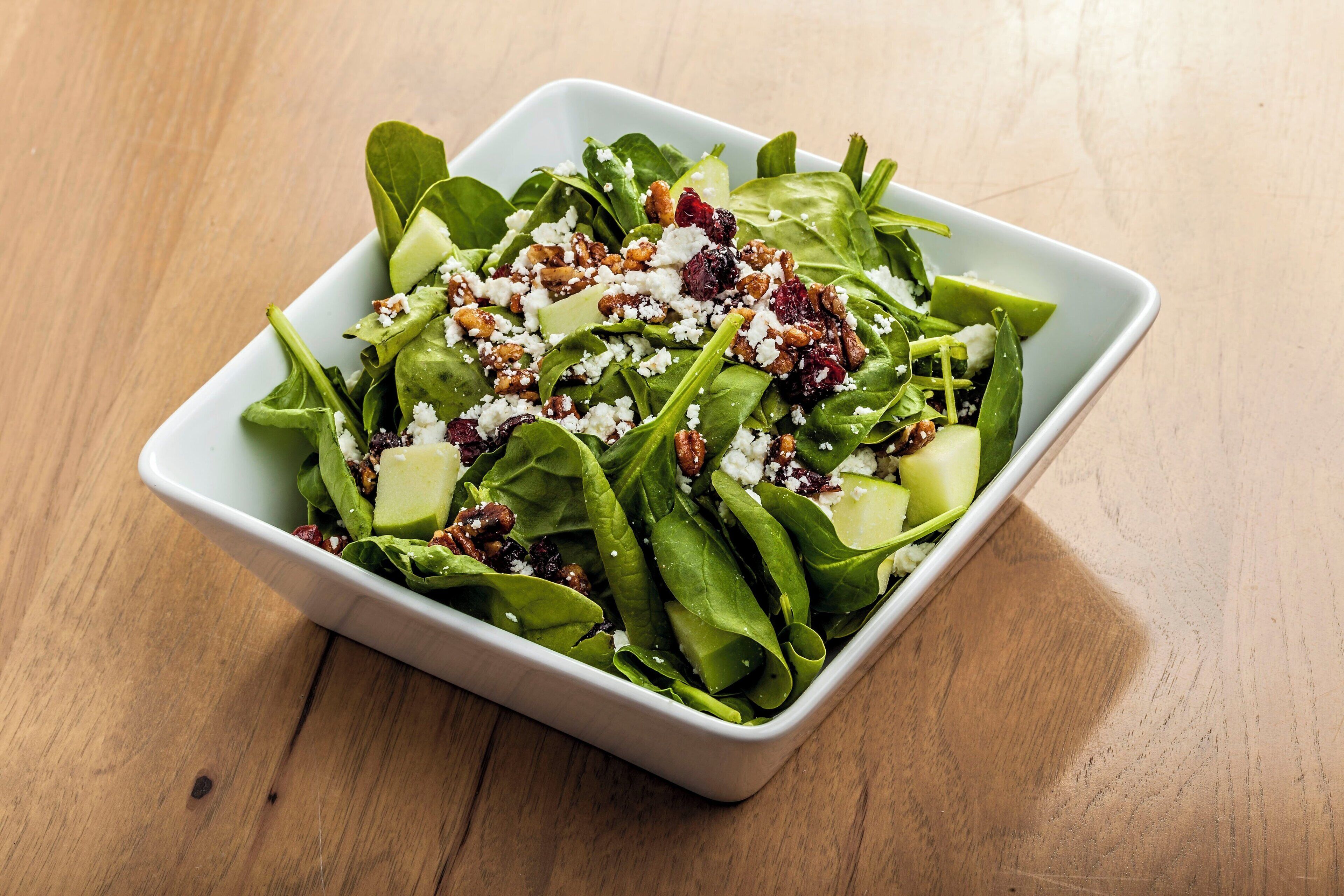 A white square bowl containing a salad with leafy greens, nuts, and crumbled cheese on a wooden table.