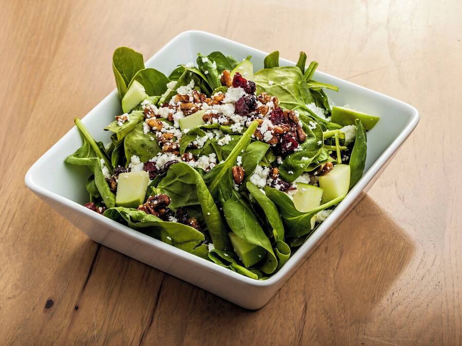 Tigela branca quadrada com salada de folhas verdes, nozes e queijo esfarelado sobre mesa de madeira.