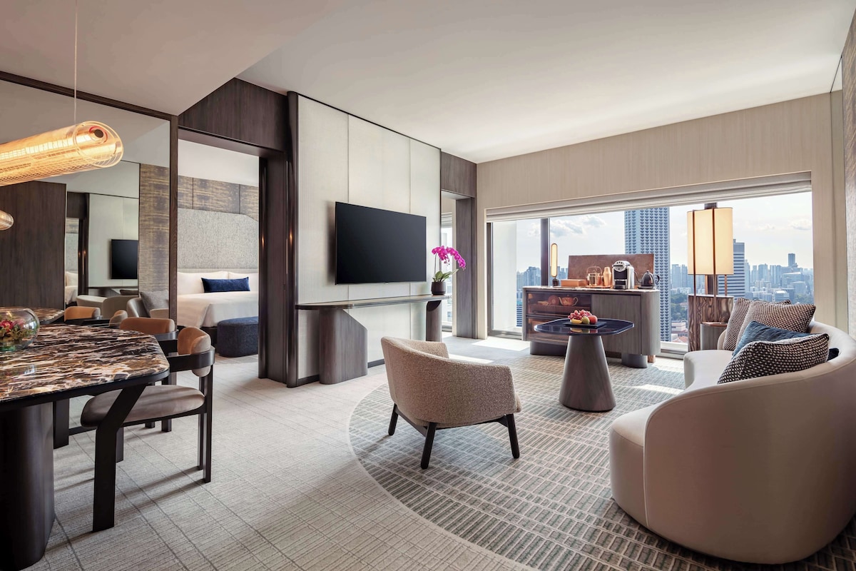 A modern hotel room with a dining area, a living room with a sofa and armchairs, a flat-screen TV, and a city view.