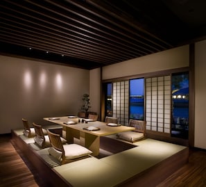 A dining area with a low table and cushioned seating, wooden beams on the ceiling, and a view through a shoji screen.