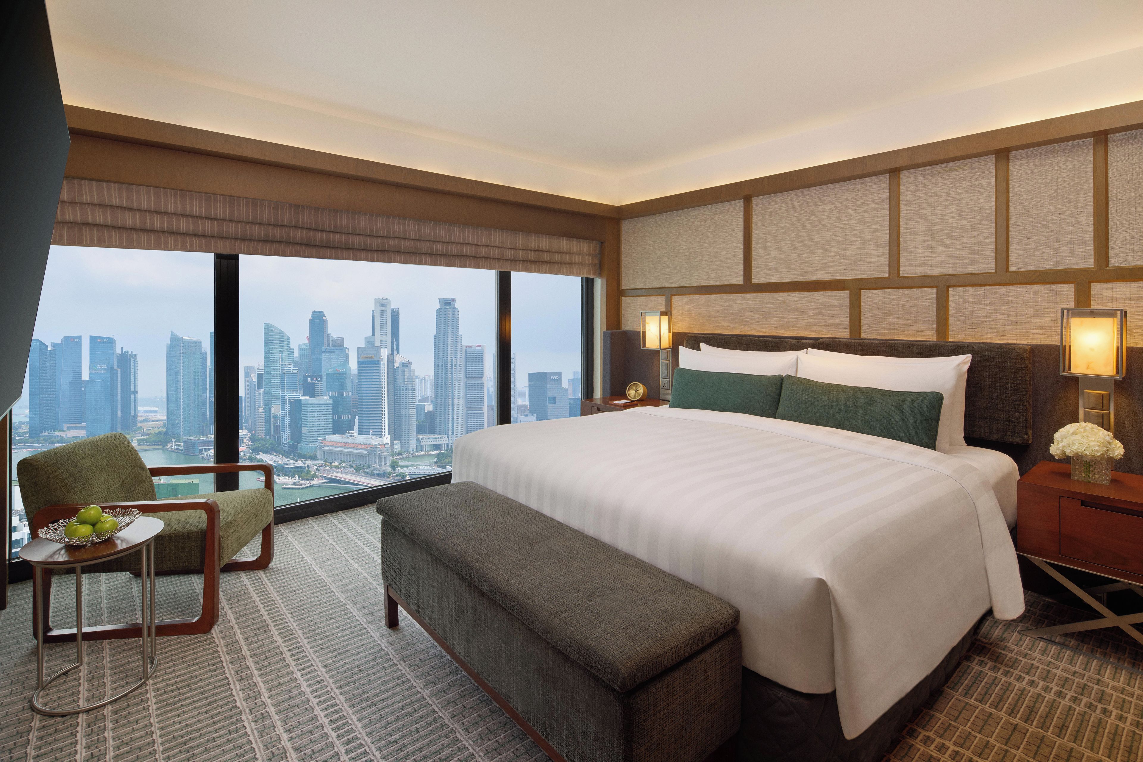 Habitación de hotel con una cama grande, una zona de estar con una silla y una mesita pequeña, una mesita de noche con lámpara y vistas a un paisaje urbano a través de amplios ventanales.