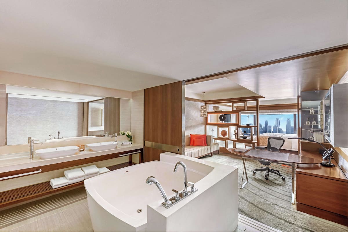 Un baño moderno con una bañera grande, dos lavamanos y un mueble de tocador. En el fondo, hay un escritorio con una silla y una estantería.