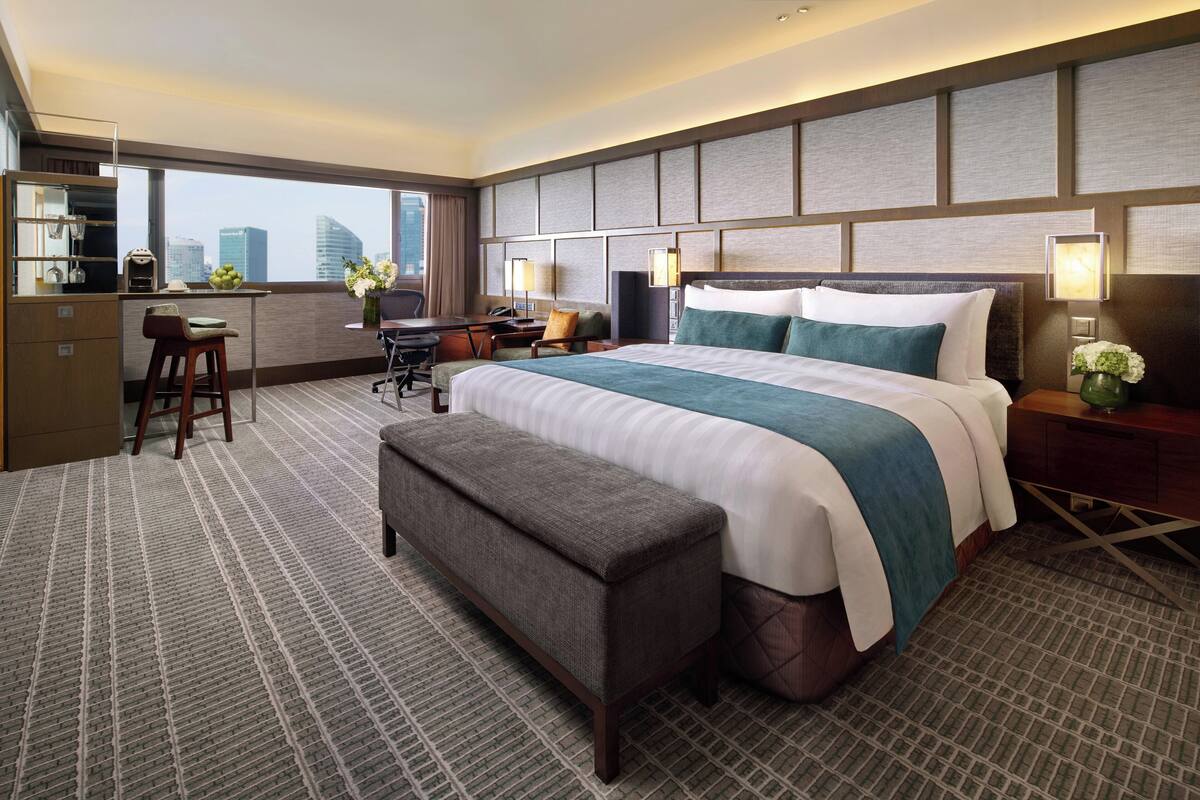 Habitación de hotel con una cama grande, un escritorio con una silla, un banco y vistas a la ciudad.