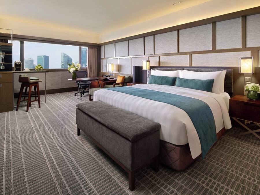 A hotel room with a large bed, a desk with a chair, a bench, and a view of the cityscape.