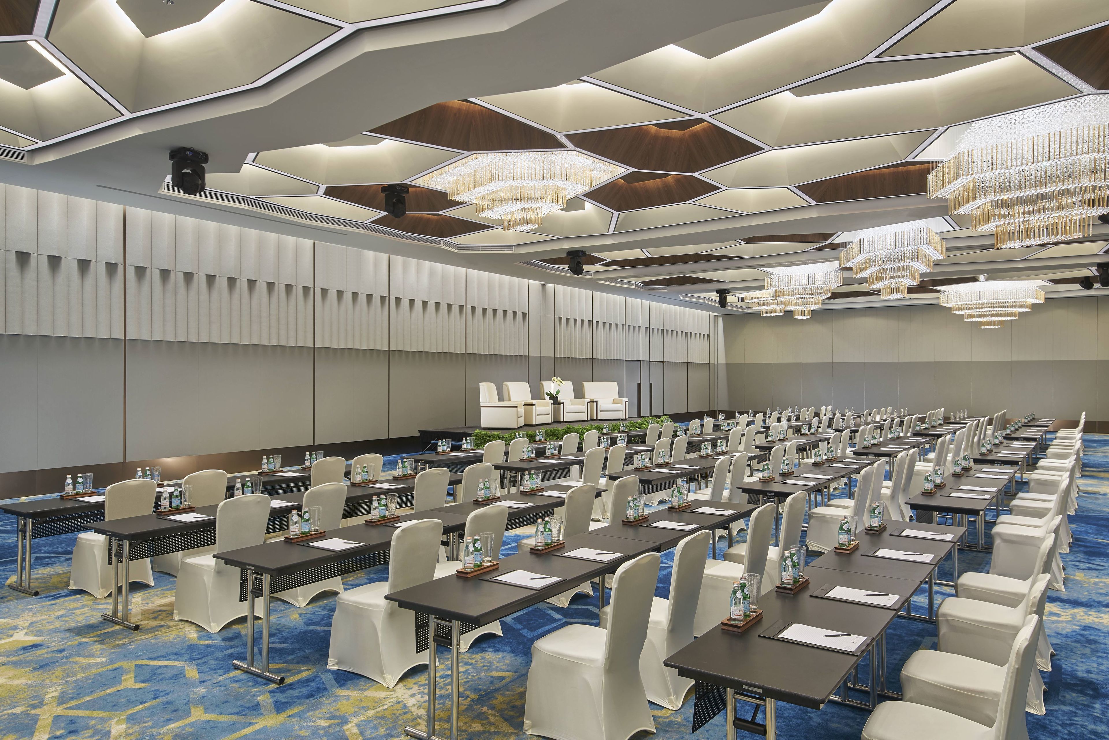 A conference room with rows of tables and chairs, a modern ceiling design, and chandeliers.