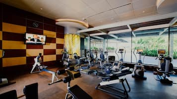 A modern gym with various exercise equipment, a television screen, and large windows offering a view of greenery.