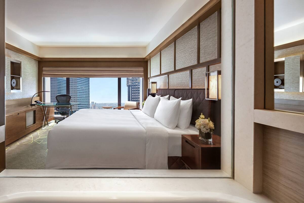 Una habitación de hotel moderna con una cama grande, un escritorio con una silla, una mesita con un jarrón de flores y vistas a edificios altos.