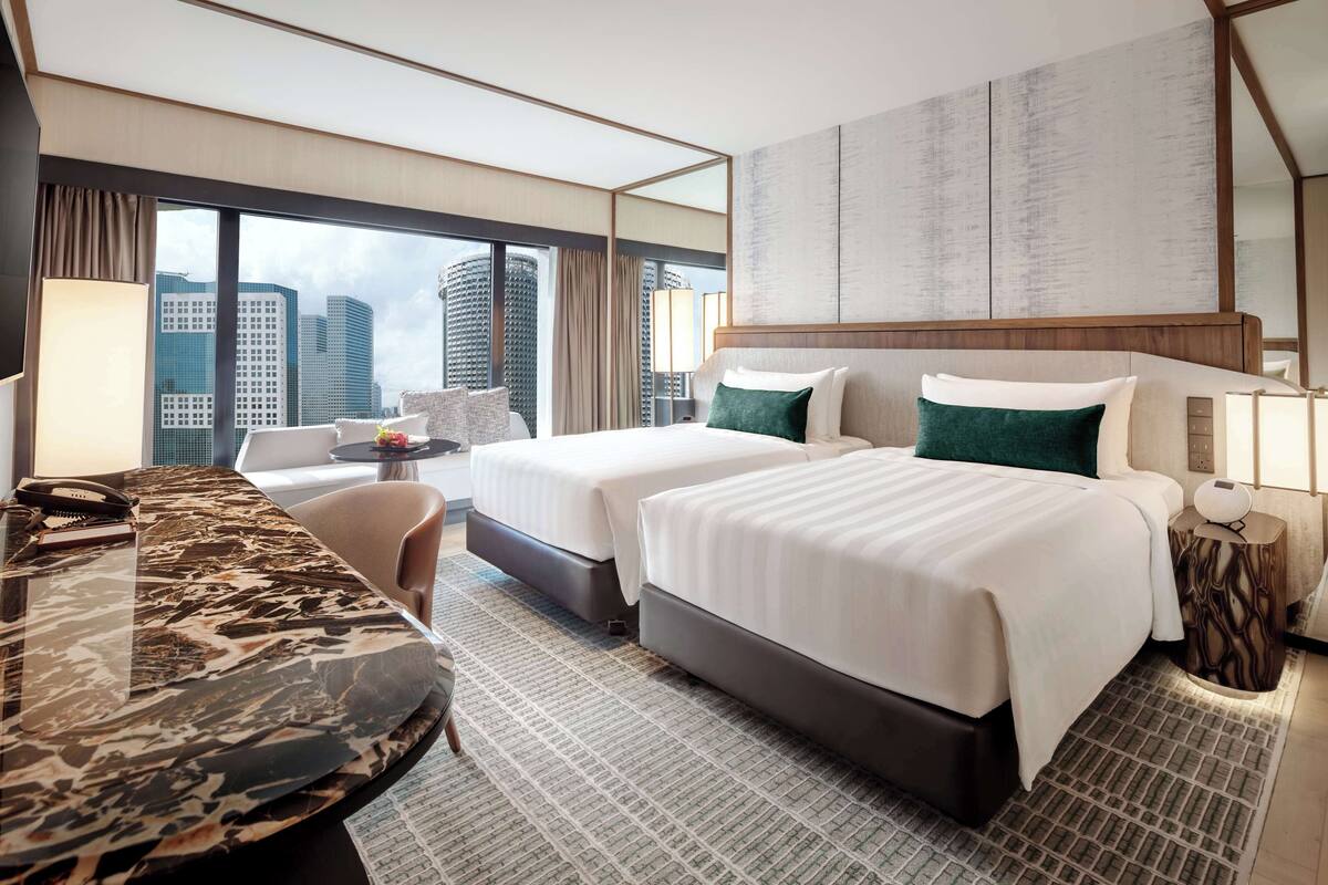 Habitación de hotel con dos camas, un escritorio y un ventanal grande con vistas a la ciudad.