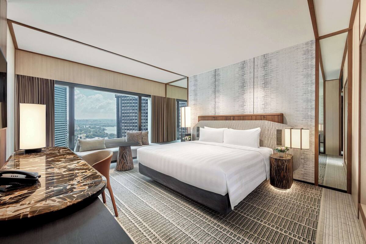 Une chambre d’hôtel moderne dotée d’un grand lit, d’un bureau avec un téléphone, d’une chaise et offrant une vue sur la ville grâce à de grandes fenêtres.