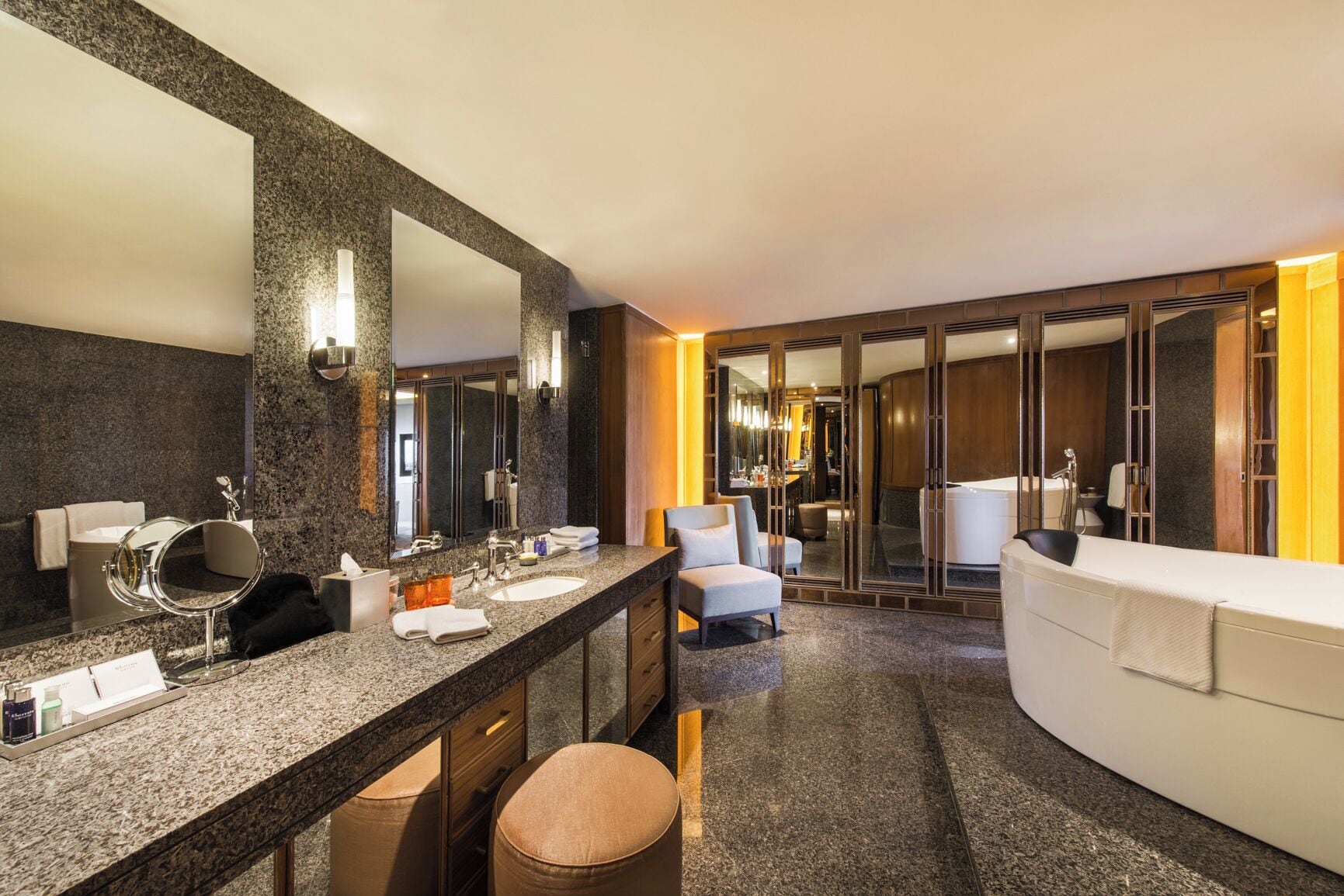 A modern bathroom with a large mirror, two sinks, a bathtub, and a seating area.