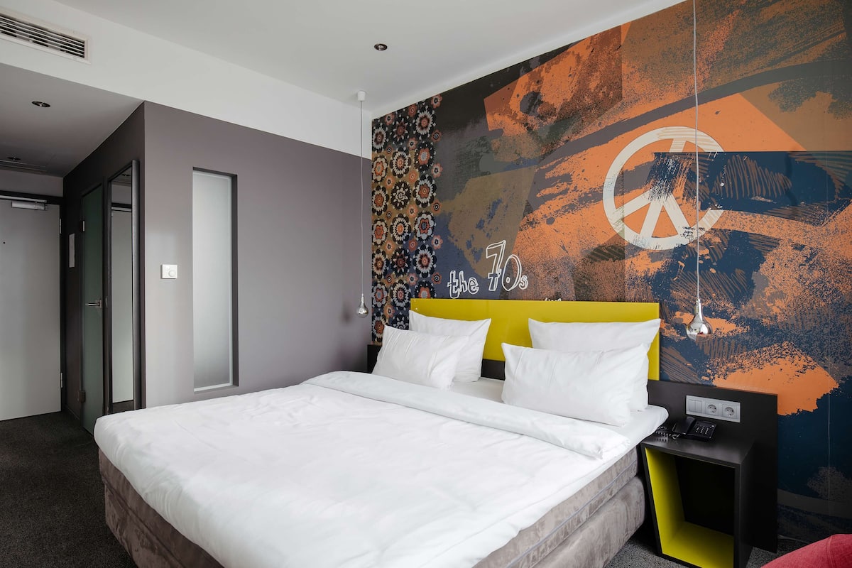 Une chambre d’hôtel moderne avec un grand lit, une tête de lit ornée d’un symbole de paix, une fresque murale et une table de chevet avec un téléphone.