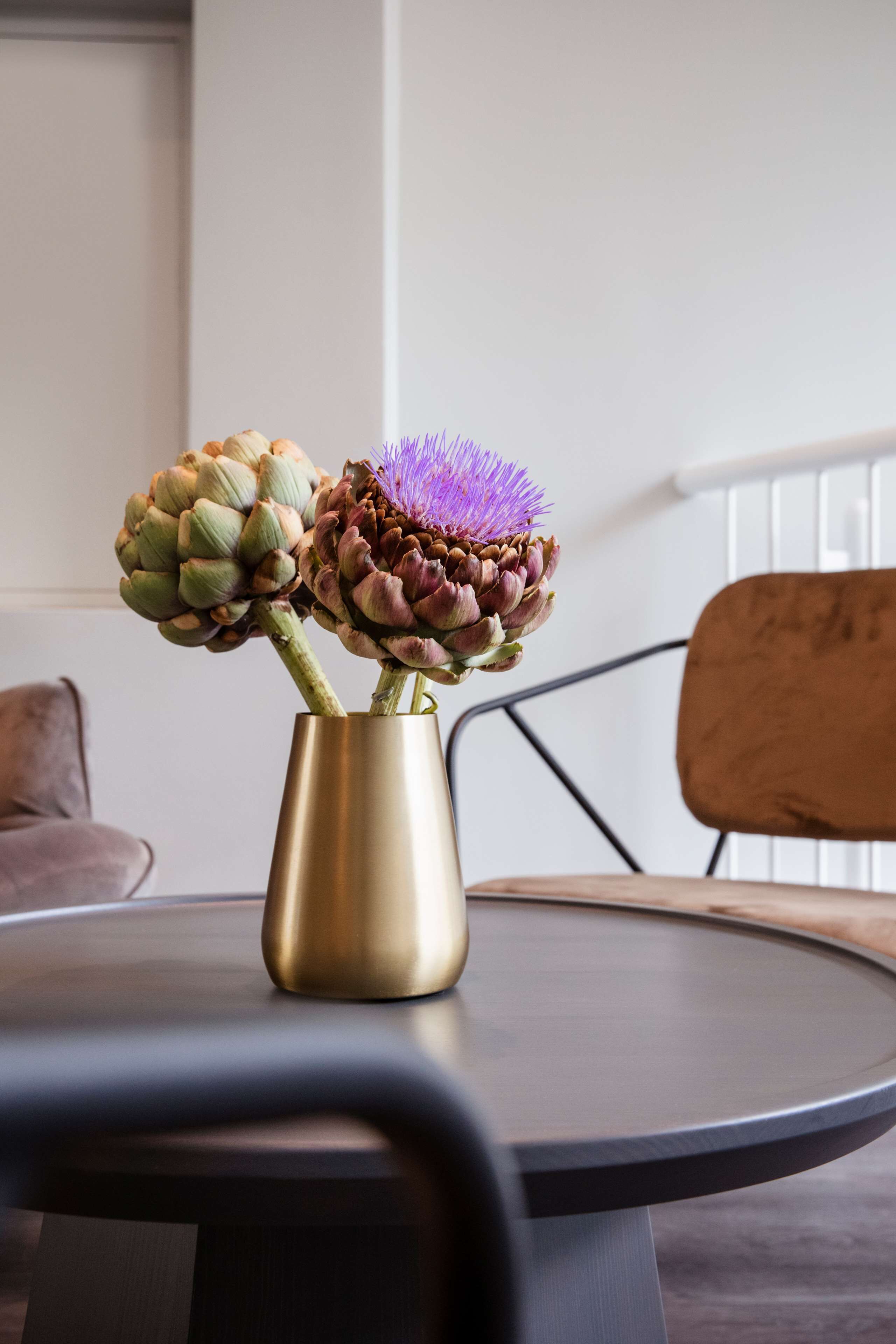 Et rundt bord med en gylden vase der indeholder to artiskokker og en lilla blomst.