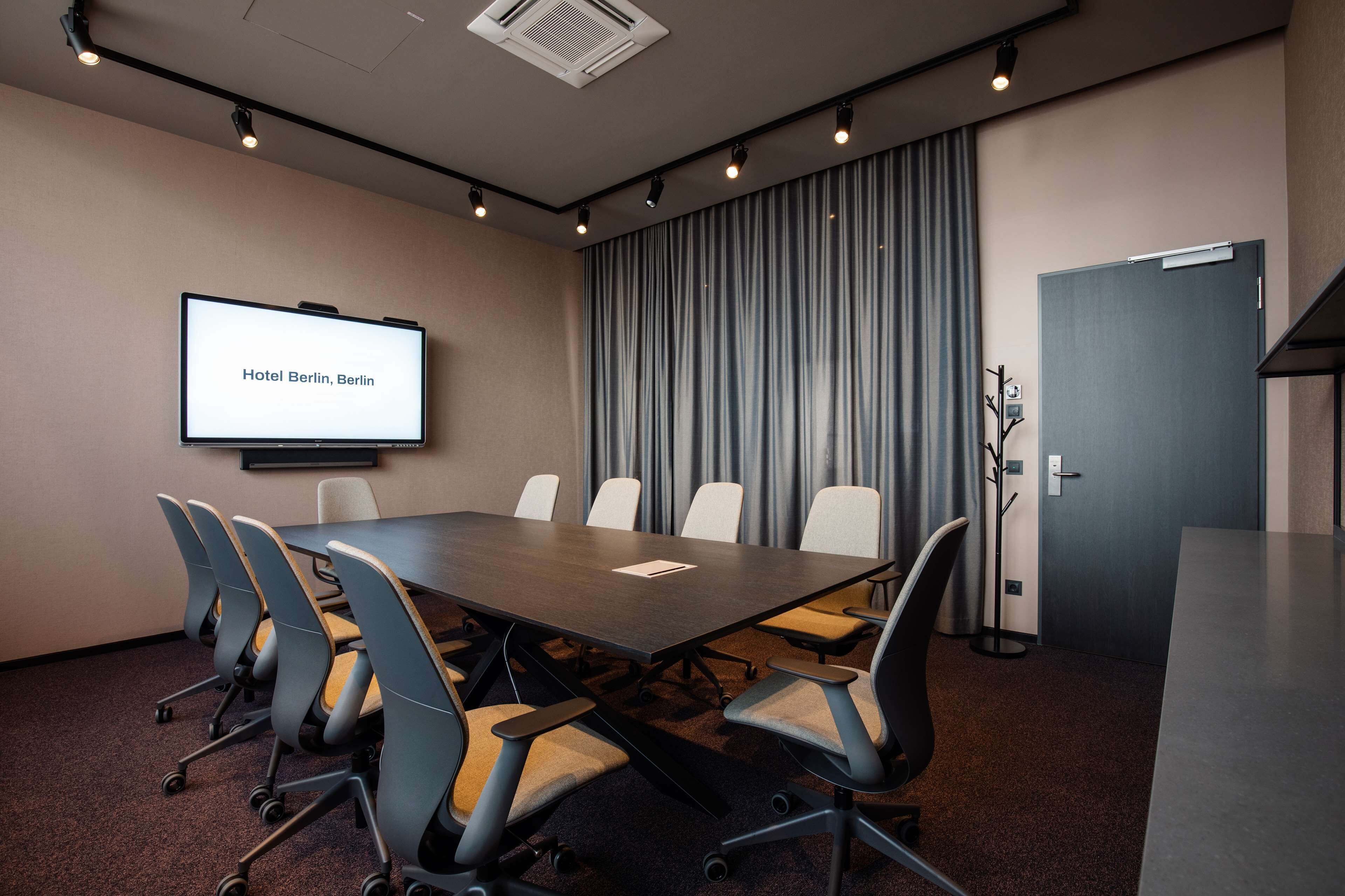 A conference room with a long table, six chairs, a projector screen displaying "Hotel Berlin, Berlin," and a door to the right.