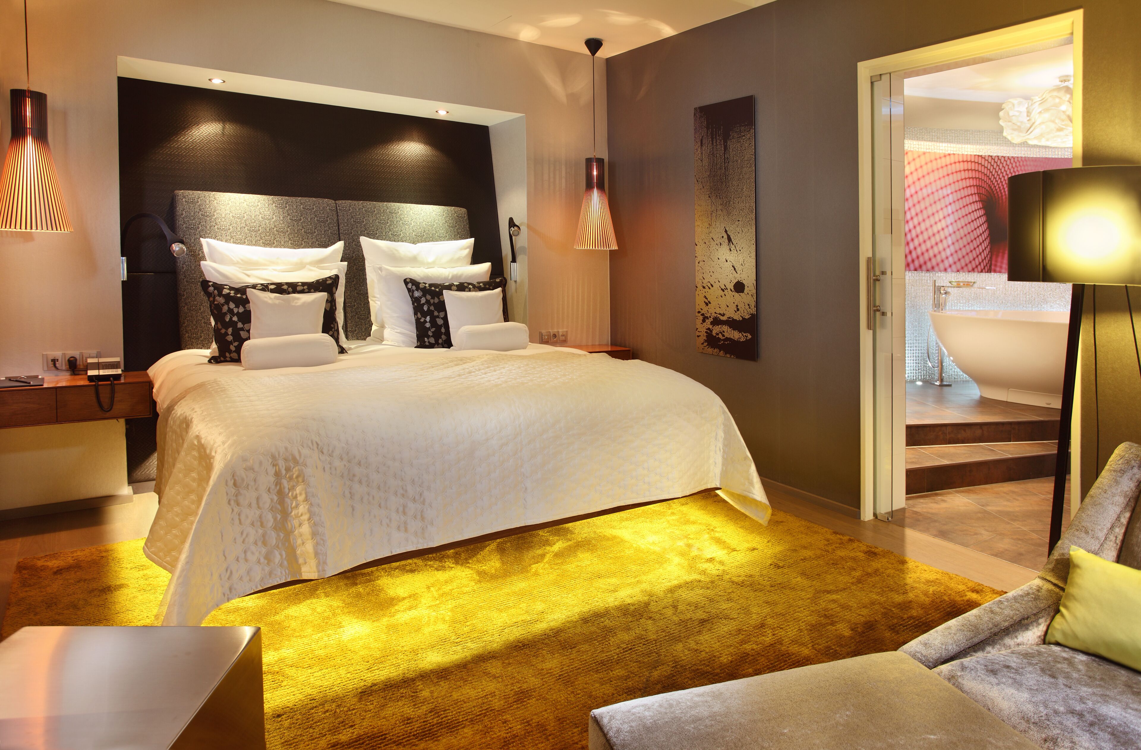 A modern hotel room with a large bed, a sofa, a desk, and a bathroom visible through an open door.
