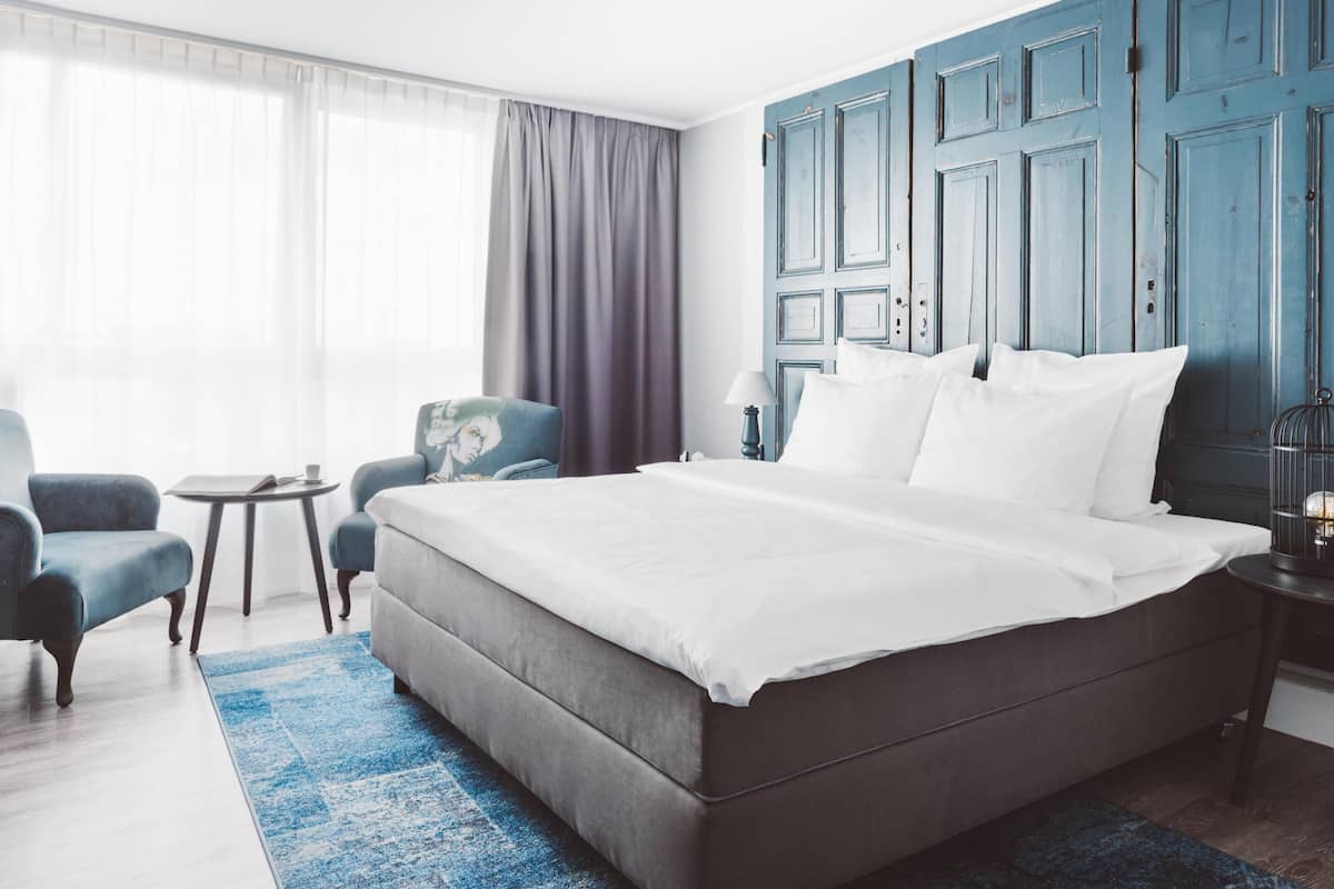 Une chambre d’hôtel avec un grand lit, une petite table, deux fauteuils, une lampe et une vue sur l’extérieur.