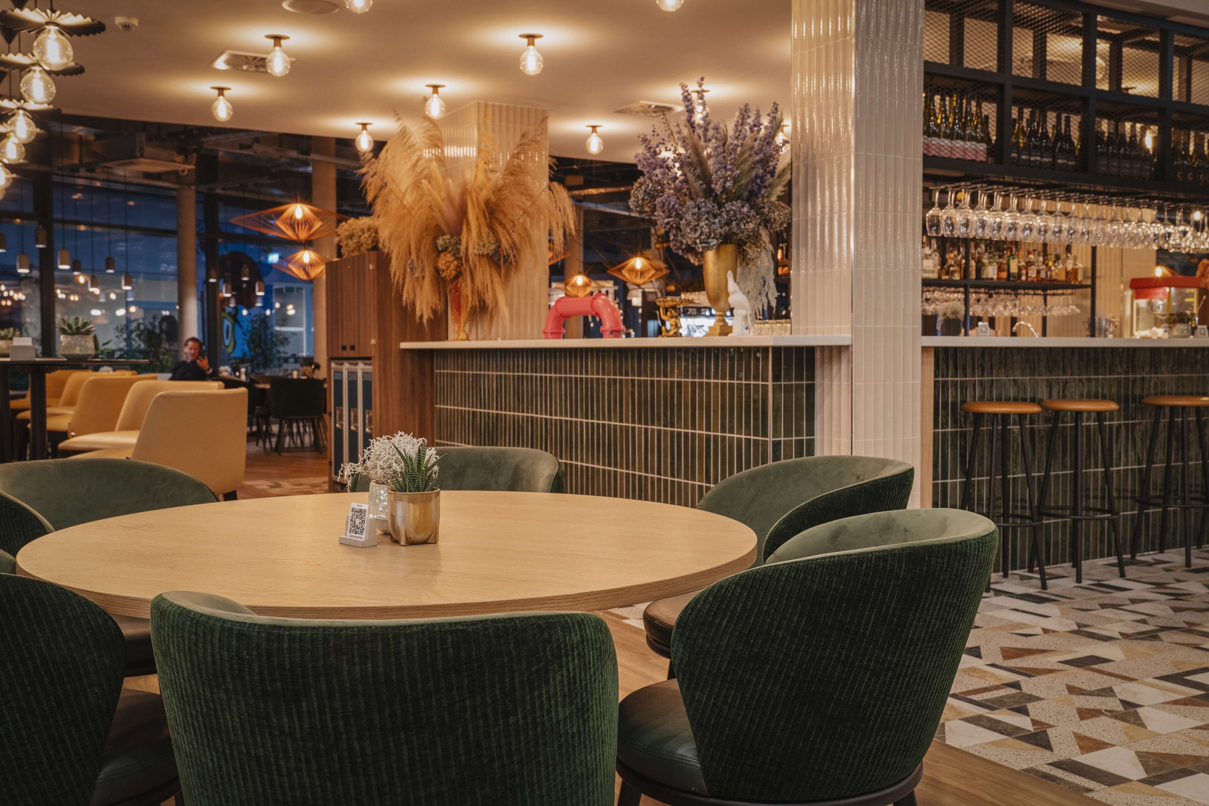 A modern bar area with a round wooden table, green upholstered chairs, and a variety of hanging lights.