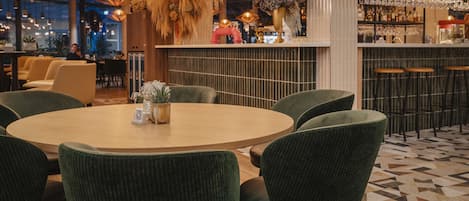 A modern bar area with a round wooden table, green upholstered chairs, and a variety of hanging lights.