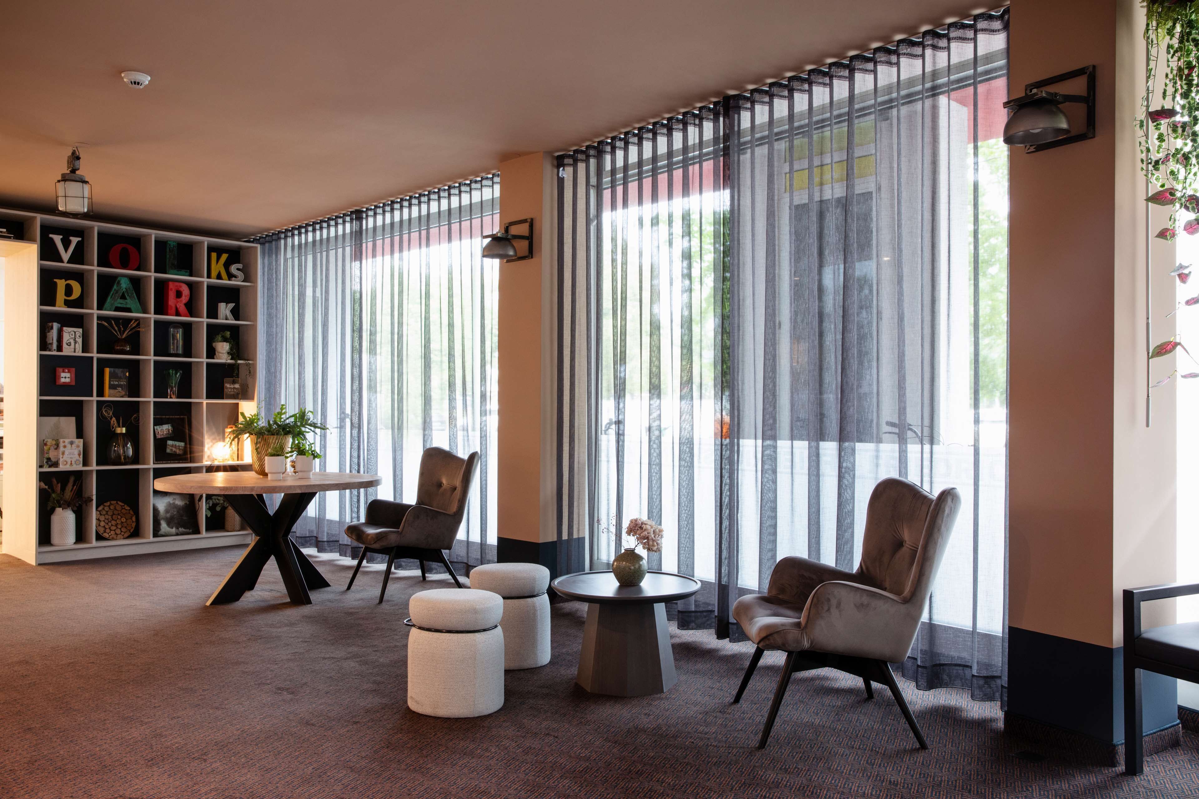Una habitación moderna con una estantería, dos sillones, una mesa redonda y un ventanal grande con persianas verticales.