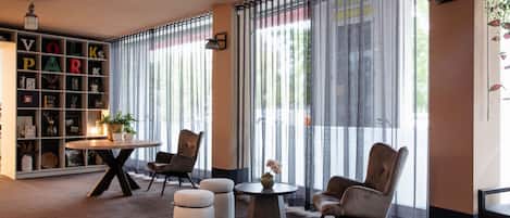 A modern room with a bookshelf, two chairs, a round table, and a large window with vertical blinds.