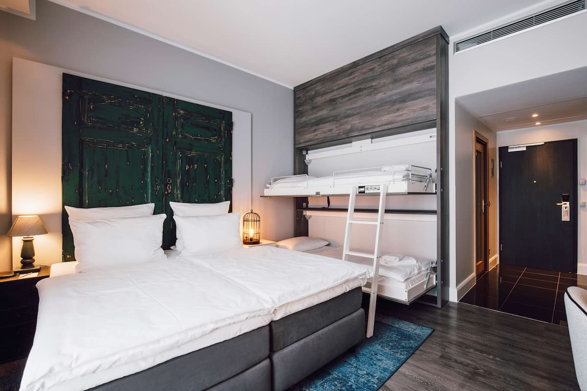 Une chambre d’hôtel moderne avec un grand lit, un lit superposé, une tête de lit en bois, une table de chevet avec une lampe et un tapis bleu.