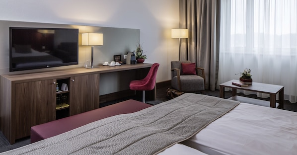 A hotel room with a flat-screen TV, a desk with a lamp, a chair, a bed, a small table, and a potted plant.