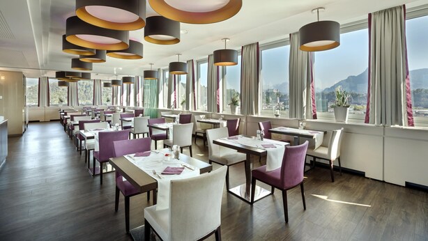 A modern restaurant with multiple dining tables and chairs, large windows with curtains, and pendant lighting. - Austria Trend Europa Salzburg (Salzburg)