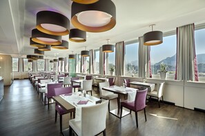 Un restaurante moderno con varias mesas y sillas, amplias ventanas con cortinas e iluminación colgante.