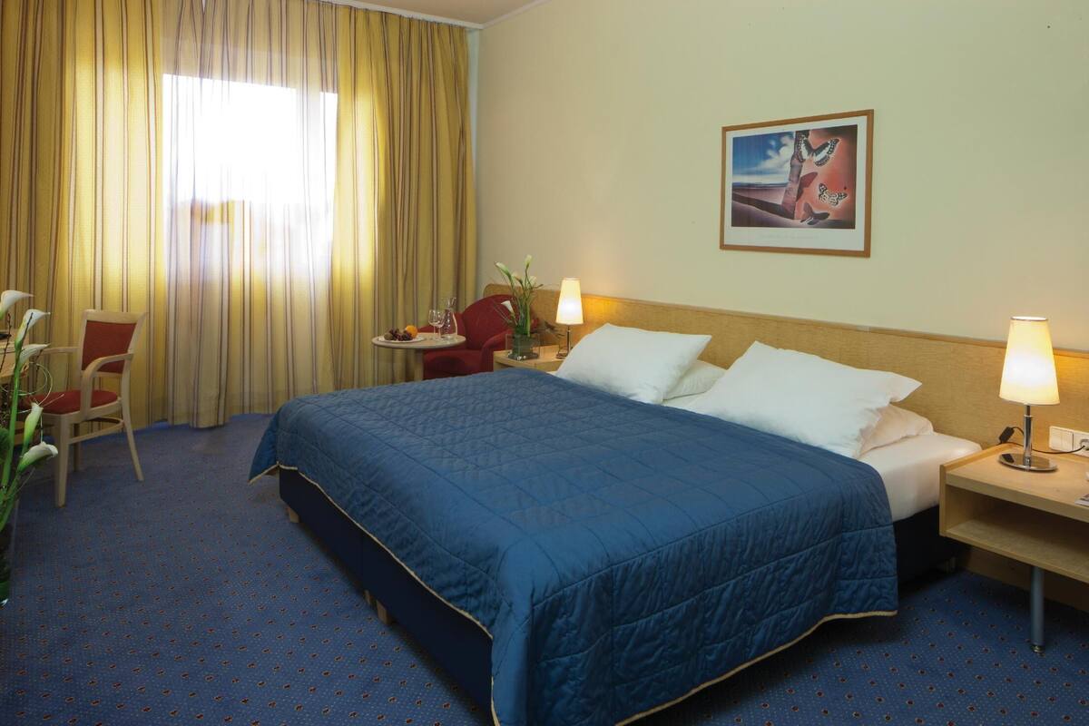 Une chambre d’hôtel avec un grand lit, deux tables de chevet avec des lampes, une petite table avec un vase et une chaise, et une fenêtre avec des rideaux.