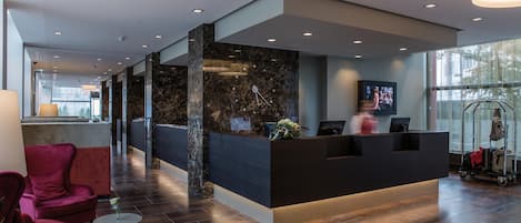 A modern reception area with a marble reception desk, a TV screen, and a luggage cart.