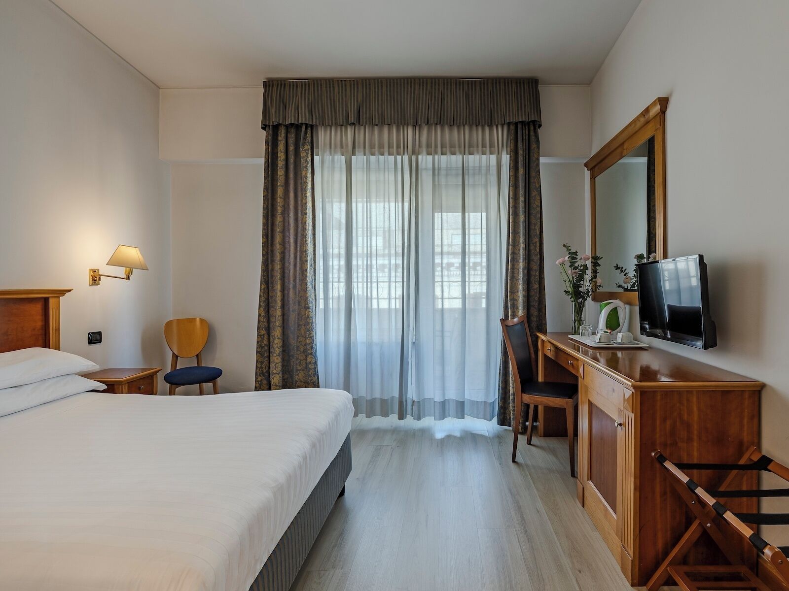 A hotel room with a bed, a desk with a chair, a television, and a window with curtains.