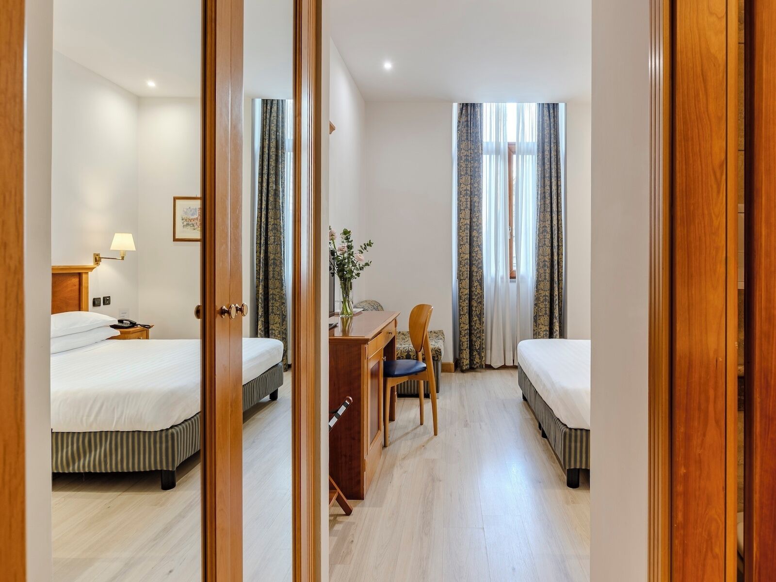 A hotel room with two beds, a desk, a chair, and a lamp.