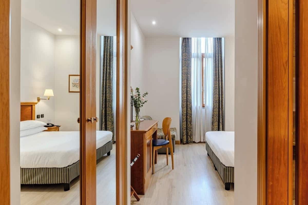A hotel room with two beds, a desk, a chair, and a lamp.