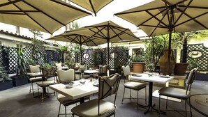 Un área de comedor al aire libre con mesas y sillas, protegida por sombrillas grandes, rodeada de plantas y una cerca decorativa.