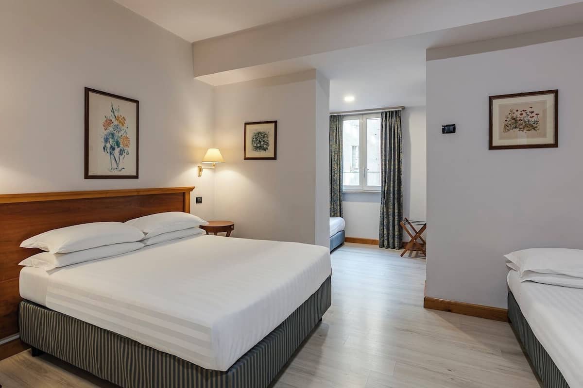Ein Hotelzimmer mit einem großen Bett, einem Nachttisch, einer Lampe, einem Fenster mit Vorhängen und einer kleinen Sitzecke.