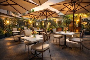 Un área de comedor al aire libre con mesas, sillas, sombrillas y plantas en macetas.