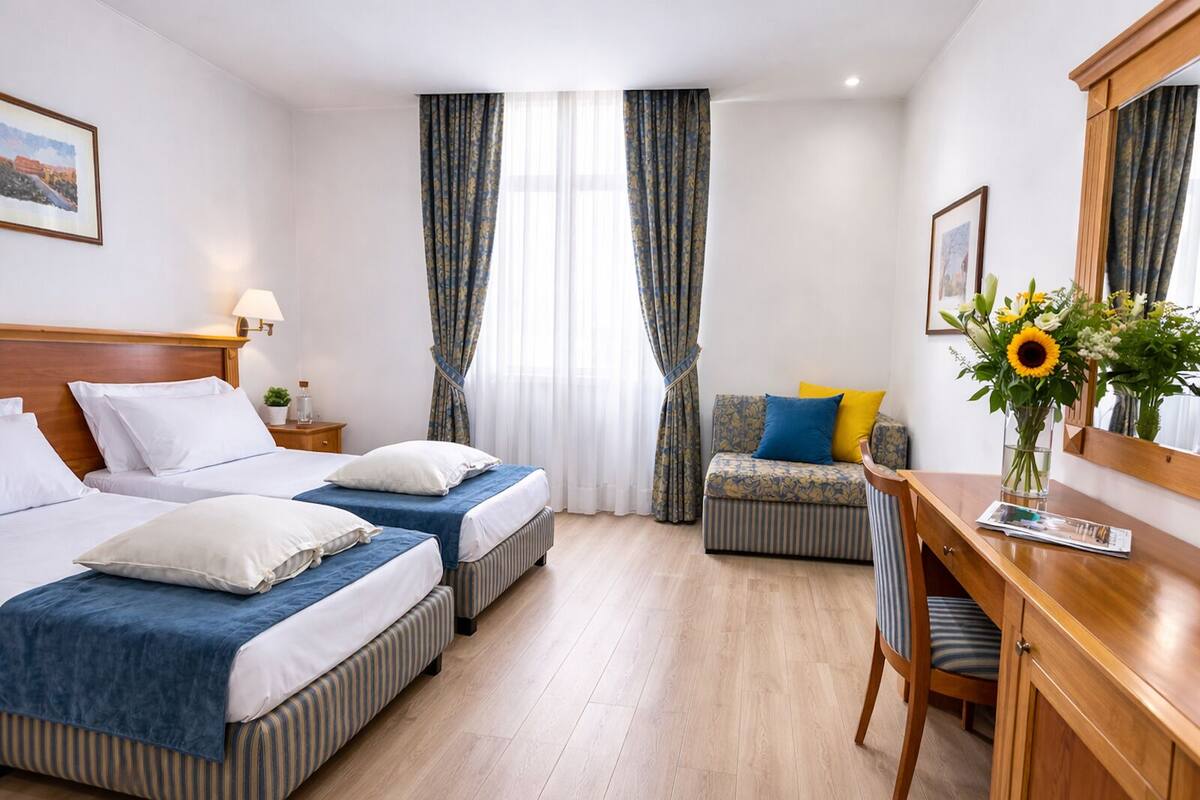 Ein Hotelzimmer mit zwei Betten, einem Schreibtisch, einem Stuhl, einer kleinen Couch, einem Spiegel und einem Fenster mit Vorhängen.